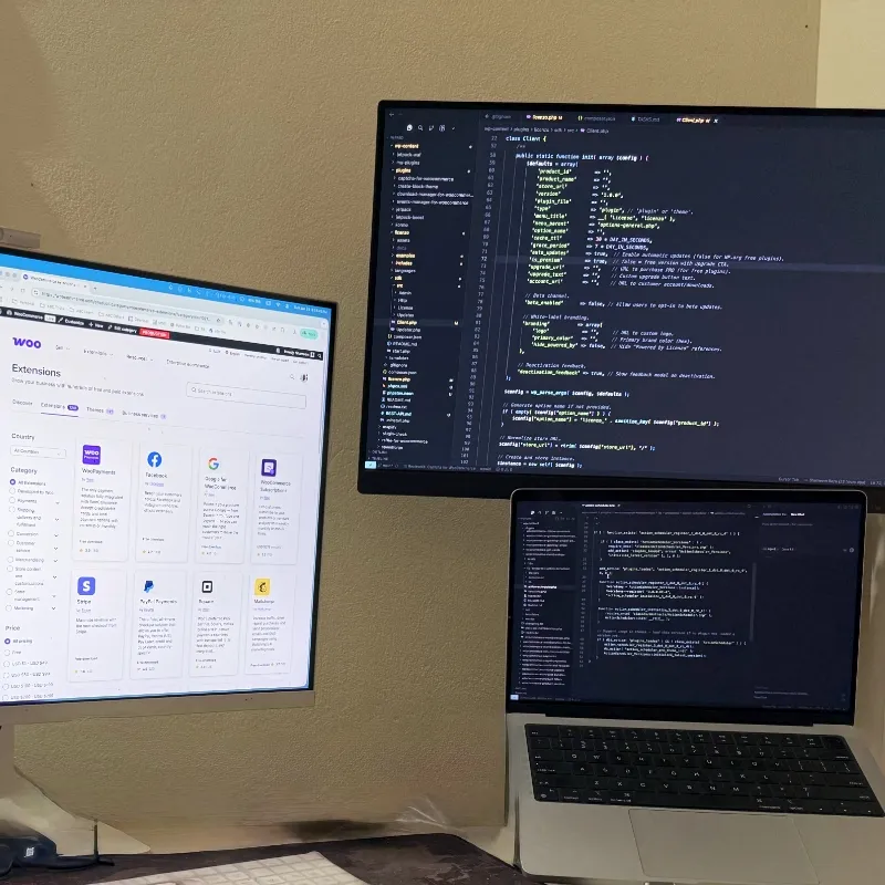 The desk: two monitors with code and a WooCommerce dashboard above a MacBook